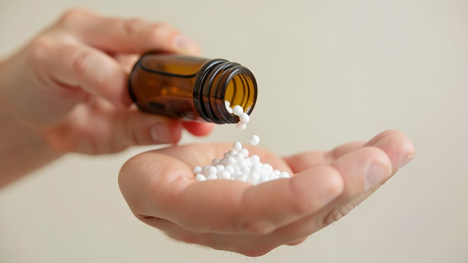 Nahaufnahme weißer Globuli, die aus einem kleinen braunen Apothekenfläschchen auf eine Handfläche rieseln